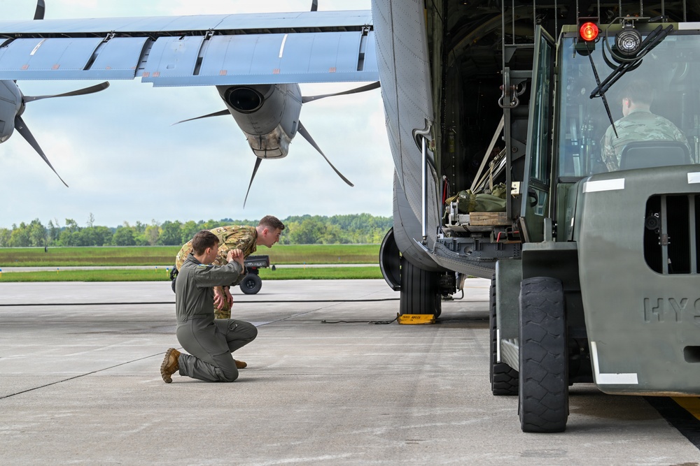 Military personnel are conducting ground operations alongside a large military transport aircraft, likely a C-130 Hercules, on a tarmac. Two service members in military uniforms are crouching and inspecting the aircraft's landing gear or undercarriage area, while a third person operates a military forklift or cargo loader positioned at the aircraft's open rear cargo ramp. The scene appears to depict a loading or pre-flight inspection operation at a military airfield.