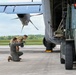 Military personnel are conducting ground operations alongside a large military transport aircraft, likely a C-130 Hercules, on a tarmac. Two service members in military uniforms are crouching and inspecting the aircraft's landing gear or undercarriage area, while a third person operates a military forklift or cargo loader positioned at the aircraft's open rear cargo ramp. The scene appears to depict a loading or pre-flight inspection operation at a military airfield.