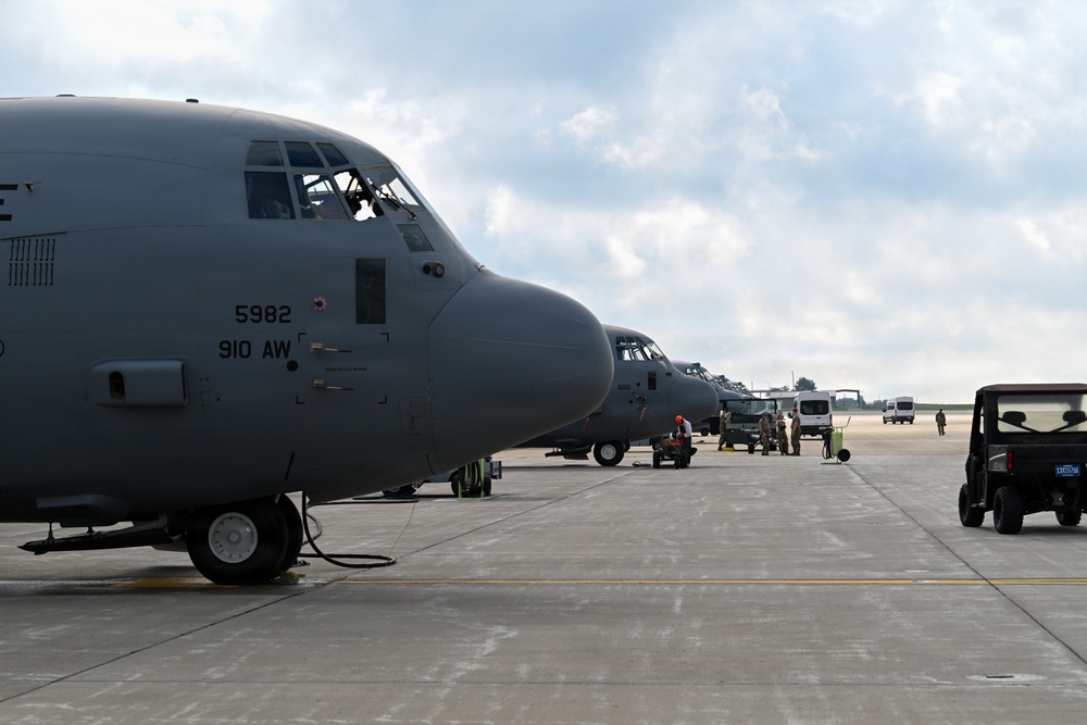 Multiple gray military C-130 Hercules transport aircraft are parked on a flight line ramp, with the closest aircraft bearing markings '5982' and '910 AW' (910th Airlift Wing). Ground crew personnel and support vehicles are visible between the aircraft performing maintenance or pre-flight operations. The scene appears to be at a military air base under partly cloudy skies.