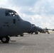 Multiple gray military C-130 Hercules transport aircraft are parked on a flight line ramp, with the closest aircraft bearing markings '5982' and '910 AW' (910th Airlift Wing). Ground crew personnel and support vehicles are visible between the aircraft performing maintenance or pre-flight operations. The scene appears to be at a military air base under partly cloudy skies.