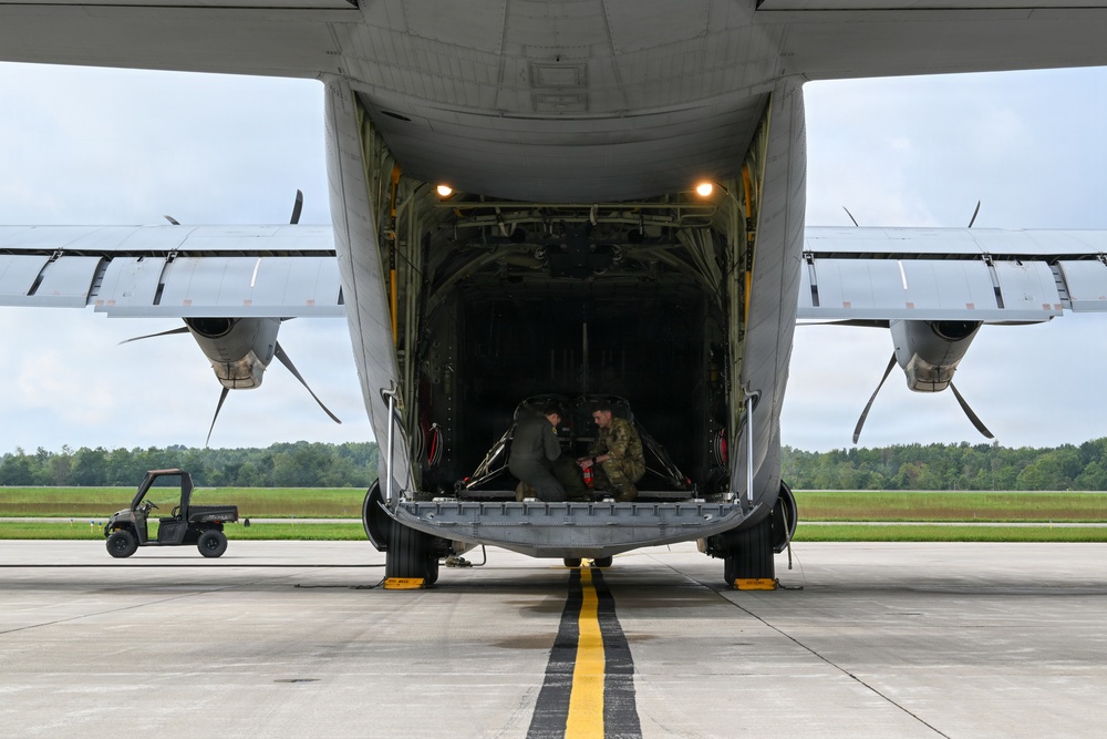 A large military cargo aircraft, consistent with a C-130 Hercules, is photographed from the rear with its loading ramp fully lowered on a concrete tarmac. Two personnel are visible inside the open cargo bay, appearing to handle equipment or cargo. A small utility vehicle is visible to the left side of the aircraft on the tarmac.