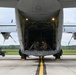 A large military cargo aircraft, consistent with a C-130 Hercules, is photographed from the rear with its loading ramp fully lowered on a concrete tarmac. Two personnel are visible inside the open cargo bay, appearing to handle equipment or cargo. A small utility vehicle is visible to the left side of the aircraft on the tarmac.