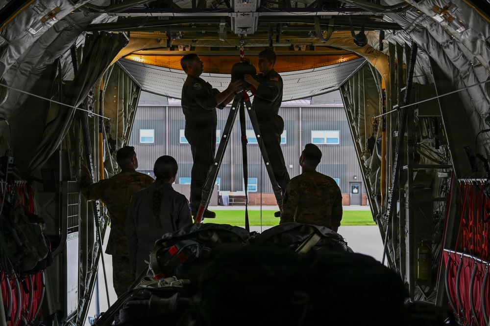 Military personnel are working inside the cargo hold of a large military transport aircraft, likely a C-130 Hercules or similar aircraft. Two individuals are standing on a ladder/stepladder working on overhead equipment near the aircraft's ceiling, while others observe or assist from below. The scene is set on a military airfield with a hangar visible in the background through the open cargo ramp.