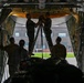 Military personnel are working inside the cargo hold of a large military transport aircraft, likely a C-130 Hercules or similar aircraft. Two individuals are standing on a ladder/stepladder working on overhead equipment near the aircraft's ceiling, while others observe or assist from below. The scene is set on a military airfield with a hangar visible in the background through the open cargo ramp.