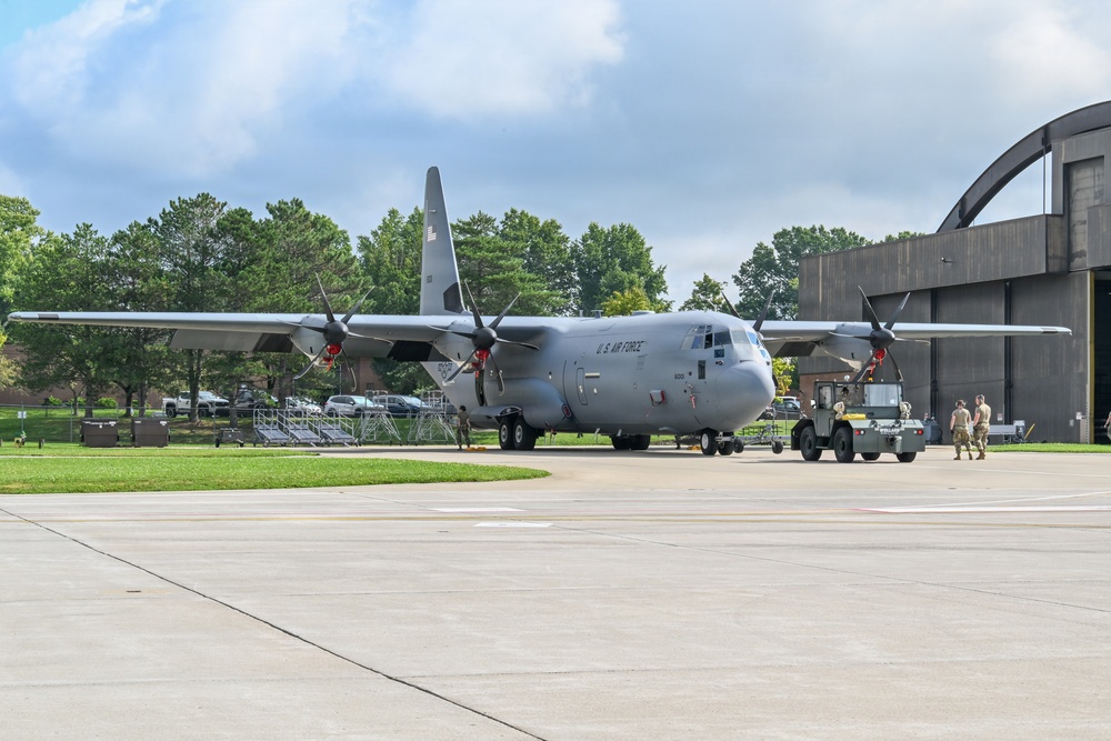 A U.S. Air Force C-130 Hercules turboprop military transport aircraft is positioned on a concrete flight line apron adjacent to a large aircraft hangar. Ground crew personnel in military uniforms are conducting towing or ground handling operations using a yellow aircraft tug vehicle. The setting appears to be an active military air base with lush green trees and partially cloudy skies in the background.