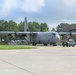 A U.S. Air Force C-130 Hercules turboprop military transport aircraft is positioned on a concrete flight line apron adjacent to a large aircraft hangar. Ground crew personnel in military uniforms are conducting towing or ground handling operations using a yellow aircraft tug vehicle. The setting appears to be an active military air base with lush green trees and partially cloudy skies in the background.