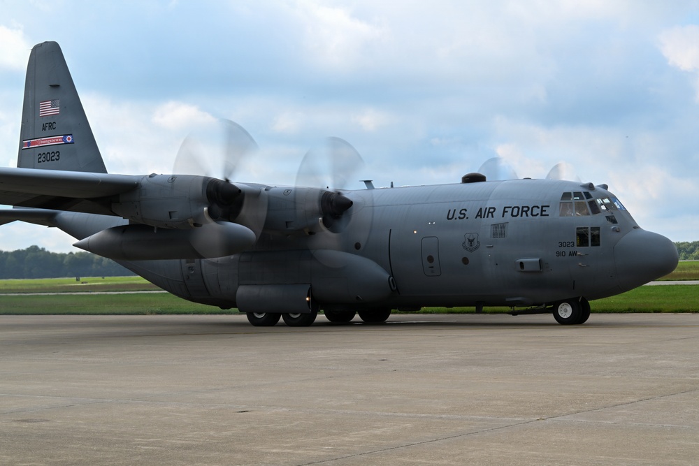 A U.S. Air Force C-130 Hercules military transport aircraft is taxiing on a concrete runway apron with all four turboprop engines running, as evidenced by the spinning propeller blur. The aircraft bears tail number 23023 and is assigned to the 910th Airlift Wing (910 AW) of the Air Force Reserve Command (AFRC). The image was taken in good lighting conditions with a partly cloudy sky and green treeline visible in the background.