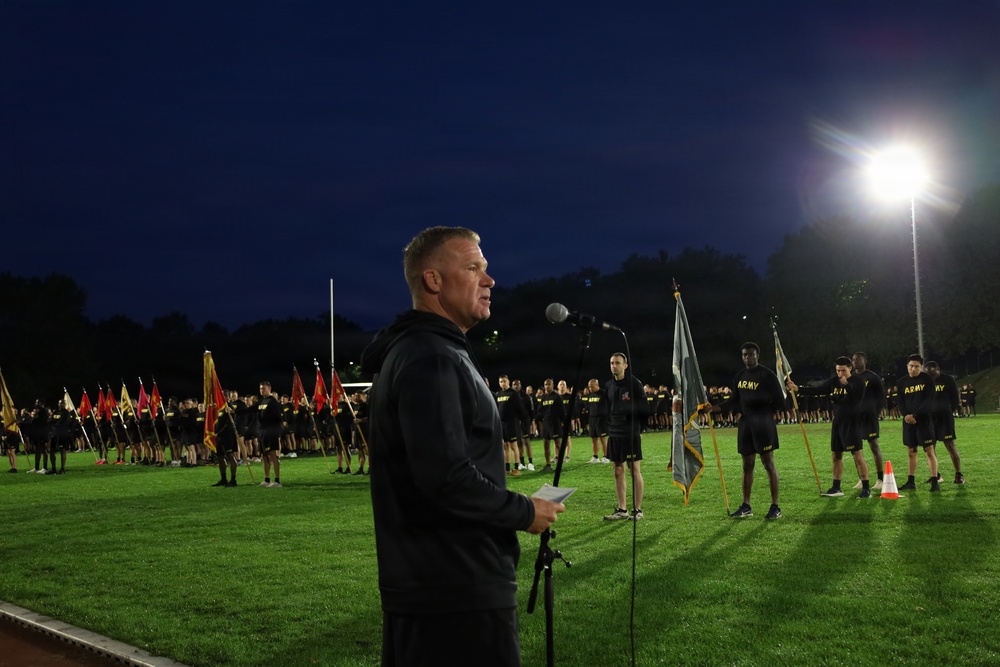 A military formation event is taking place on a lit athletic field at night or early morning. A civilian or officer in a dark tracksuit addresses a large group of personnel wearing black Army PT (physical training) uniforms, with color guard bearers holding unit guidons and an Army flag visible in the background. The scene appears to be a formal physical training formation or ceremony.