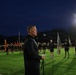 A military formation event is taking place on a lit athletic field at night or early morning. A civilian or officer in a dark tracksuit addresses a large group of personnel wearing black Army PT (physical training) uniforms, with color guard bearers holding unit guidons and an Army flag visible in the background. The scene appears to be a formal physical training formation or ceremony.