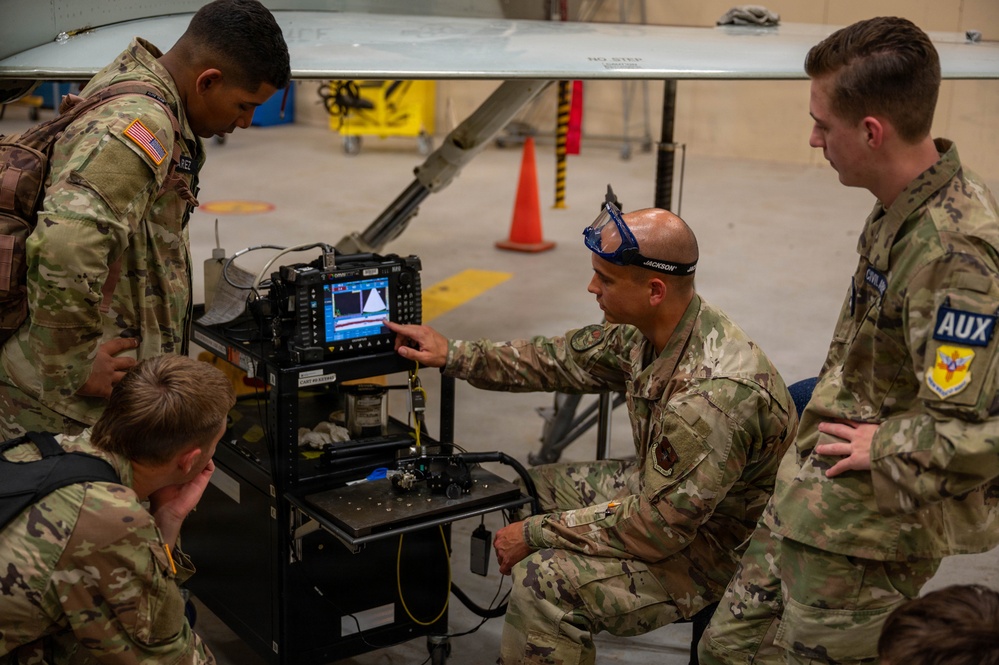 Military personnel in OCP (Operational Camouflage Pattern) uniforms are gathered around a diagnostic or testing equipment cart in what appears to be an aircraft hangar. One individual, identified by a name tape as 'Jackson,' is pointing at a monitor displaying waveform or spectrum data while instructing others. A large aircraft wing with a 'NO STEP' marking is visible in the background.