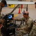 Military personnel in OCP (Operational Camouflage Pattern) uniforms are gathered around a diagnostic or testing equipment cart in what appears to be an aircraft hangar. One individual, identified by a name tape as 'Jackson,' is pointing at a monitor displaying waveform or spectrum data while instructing others. A large aircraft wing with a 'NO STEP' marking is visible in the background.