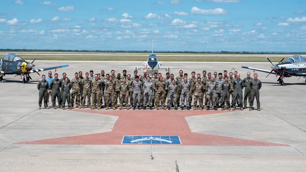 A large group of U.S. Air Force military personnel pose for a formal group photograph on an airfield tarmac. Three aircraft are visible behind them — a propeller-driven trainer on the left, a jet trainer in the center, and another propeller aircraft on the right marked 'U.S. AIR FORCE'. The personnel are wearing a mix of flight suits and camouflage uniforms, standing on a painted star emblem on the concrete apron.