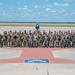 A large group of U.S. Air Force military personnel pose for a formal group photograph on an airfield tarmac. Three aircraft are visible behind them — a propeller-driven trainer on the left, a jet trainer in the center, and another propeller aircraft on the right marked 'U.S. AIR FORCE'. The personnel are wearing a mix of flight suits and camouflage uniforms, standing on a painted star emblem on the concrete apron.