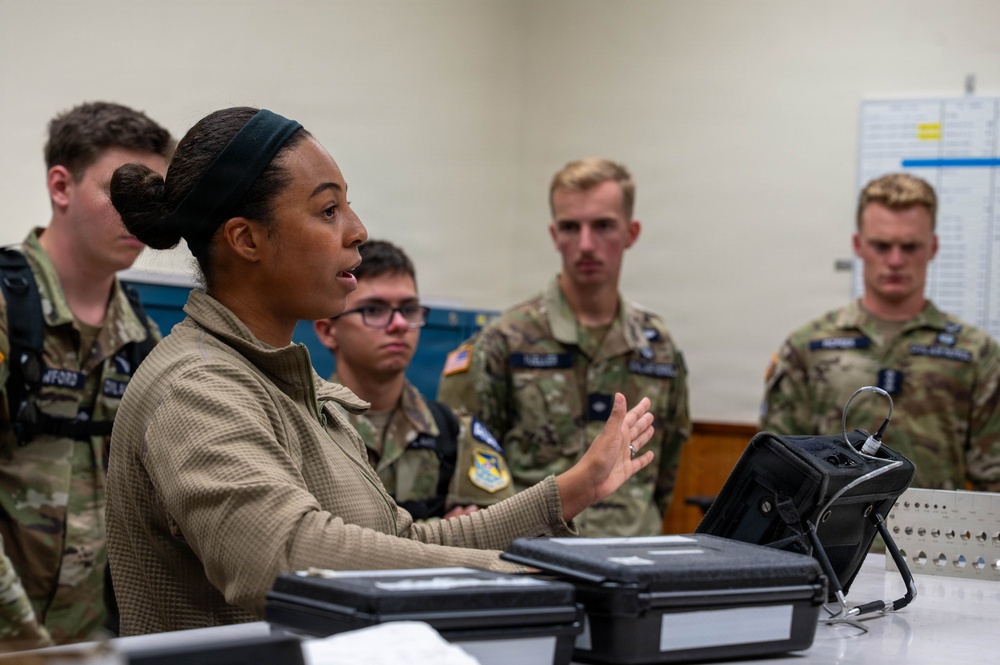 A female military instructor in a tan/olive uniform is briefing or instructing a group of younger military personnel wearing MultiCam/OCP camouflage uniforms. The setting appears to be an indoor training or operations room with equipment cases and a tablet on the table in front of her. The students are attentively listening as she gestures and speaks.
