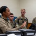 A female military instructor in a tan/olive uniform is briefing or instructing a group of younger military personnel wearing MultiCam/OCP camouflage uniforms. The setting appears to be an indoor training or operations room with equipment cases and a tablet on the table in front of her. The students are attentively listening as she gestures and speaks.
