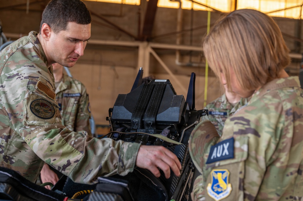 Two military personnel in OCP (Operational Camouflage Pattern) uniforms are working together on a piece of equipment inside what appears to be a hangar or maintenance facility. The individual on the right wears an 'AUX' patch and a Civil Air Patrol emblem, indicating auxiliary/CAP affiliation. They appear to be conducting hands-on training or maintenance on a military avionics or electronic device.