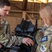 Two military personnel in OCP (Operational Camouflage Pattern) uniforms are working together on a piece of equipment inside what appears to be a hangar or maintenance facility. The individual on the right wears an 'AUX' patch and a Civil Air Patrol emblem, indicating auxiliary/CAP affiliation. They appear to be conducting hands-on training or maintenance on a military avionics or electronic device.