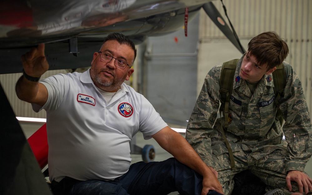 A man named Joe Benavidez, wearing a white Civil Air Patrol polo shirt, appears to be instructing or explaining something about an aircraft to a young person in Civil Air Patrol cadet uniform. They are positioned underneath or beside an aircraft, likely in a hangar setting. The interaction suggests a training or orientation session related to aircraft inspection or maintenance.