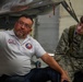 A man named Joe Benavidez, wearing a white Civil Air Patrol polo shirt, appears to be instructing or explaining something about an aircraft to a young person in Civil Air Patrol cadet uniform. They are positioned underneath or beside an aircraft, likely in a hangar setting. The interaction suggests a training or orientation session related to aircraft inspection or maintenance.