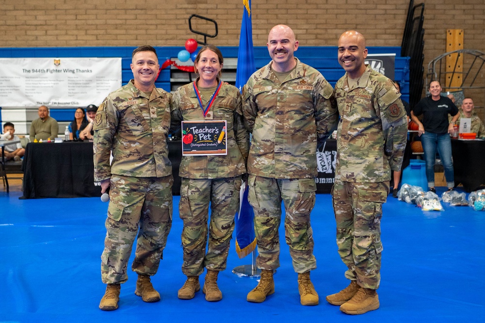 Four U.S. Air Force personnel in OCP (Operational Camouflage Pattern) uniforms pose together at what appears to be an indoor military event or ceremony. One female service member is holding a 'Teacher's Pet - Distinguished Graduate' award placard and wearing a medal around her neck, indicating she is being recognized for academic achievement. The event appears to be hosted by the 944th Fighter Wing, as indicated by a banner visible in the background.