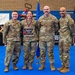 Four U.S. Air Force personnel in OCP (Operational Camouflage Pattern) uniforms pose together at what appears to be an indoor military event or ceremony. One female service member is holding a 'Teacher's Pet - Distinguished Graduate' award placard and wearing a medal around her neck, indicating she is being recognized for academic achievement. The event appears to be hosted by the 944th Fighter Wing, as indicated by a banner visible in the background.