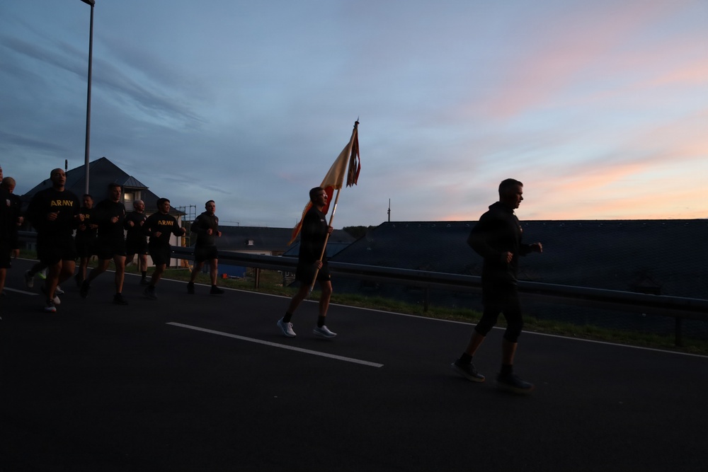 A group of individuals wearing black 'ARMY' branded athletic clothing are running along a road at dawn or dusk. One runner in the center carries a military guidon or unit flag. The scene appears to be a military physical training formation run conducted in low-light conditions.