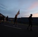 A group of individuals wearing black 'ARMY' branded athletic clothing are running along a road at dawn or dusk. One runner in the center carries a military guidon or unit flag. The scene appears to be a military physical training formation run conducted in low-light conditions.