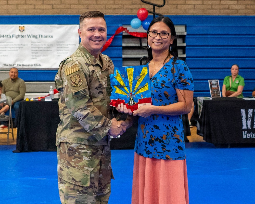 A military service member in Army Combat Uniform (ACU/OCP camouflage) is shaking hands with a civilian woman while presenting her with an award or plaque. The event appears to be a recognition ceremony hosted by the 944th Fighter Wing, taking place in a gymnasium. A banner in the background reads '944th Fighter Wing Thanks' and '944th FW Booster Club.'