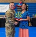 A military service member in Army Combat Uniform (ACU/OCP camouflage) is shaking hands with a civilian woman while presenting her with an award or plaque. The event appears to be a recognition ceremony hosted by the 944th Fighter Wing, taking place in a gymnasium. A banner in the background reads '944th Fighter Wing Thanks' and '944th FW Booster Club.'