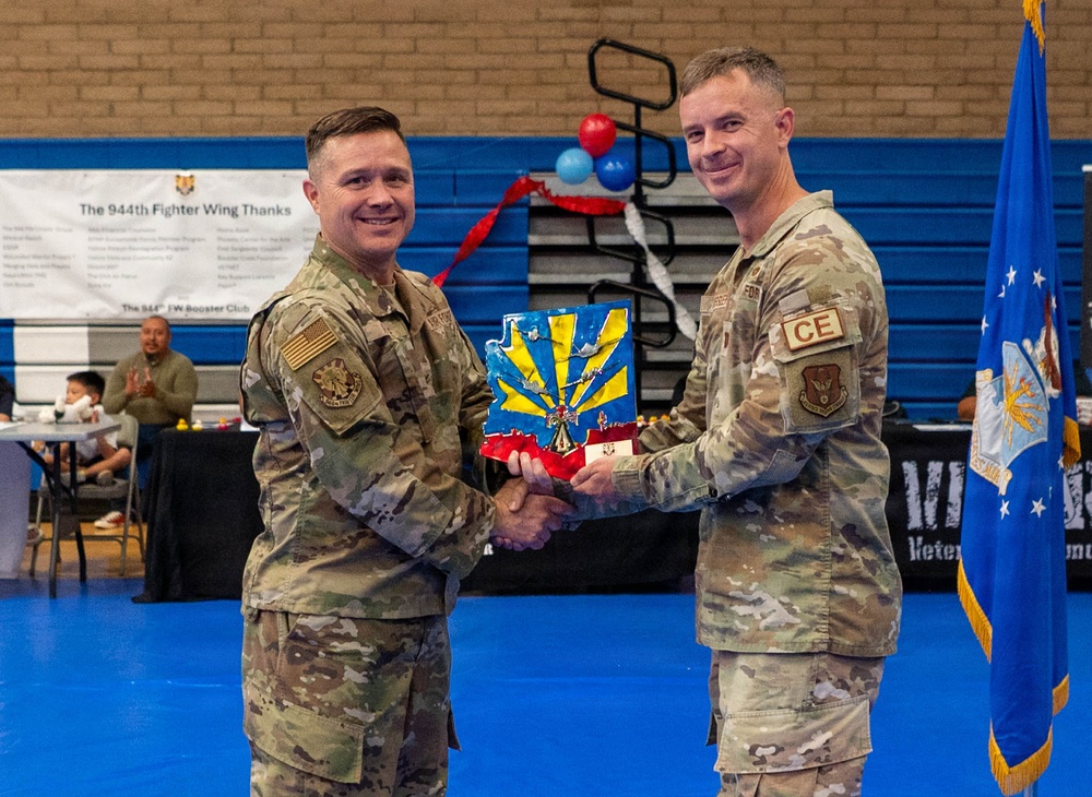 Two U.S. Air Force personnel in OCP (Operational Camouflage Pattern) uniforms are shaking hands while jointly holding a decorative plaque or award at what appears to be a 944th Fighter Wing event. The ceremony is taking place inside a gymnasium with blue floors, and a banner in the background reads 'The 944th Fighter Wing Thanks.' The individual on the right has a 'CE' (Civil Engineer) tab visible on their uniform.