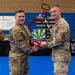 Two U.S. Air Force personnel in OCP (Operational Camouflage Pattern) uniforms are shaking hands while jointly holding a decorative plaque or award at what appears to be a 944th Fighter Wing event. The ceremony is taking place inside a gymnasium with blue floors, and a banner in the background reads 'The 944th Fighter Wing Thanks.' The individual on the right has a 'CE' (Civil Engineer) tab visible on their uniform.