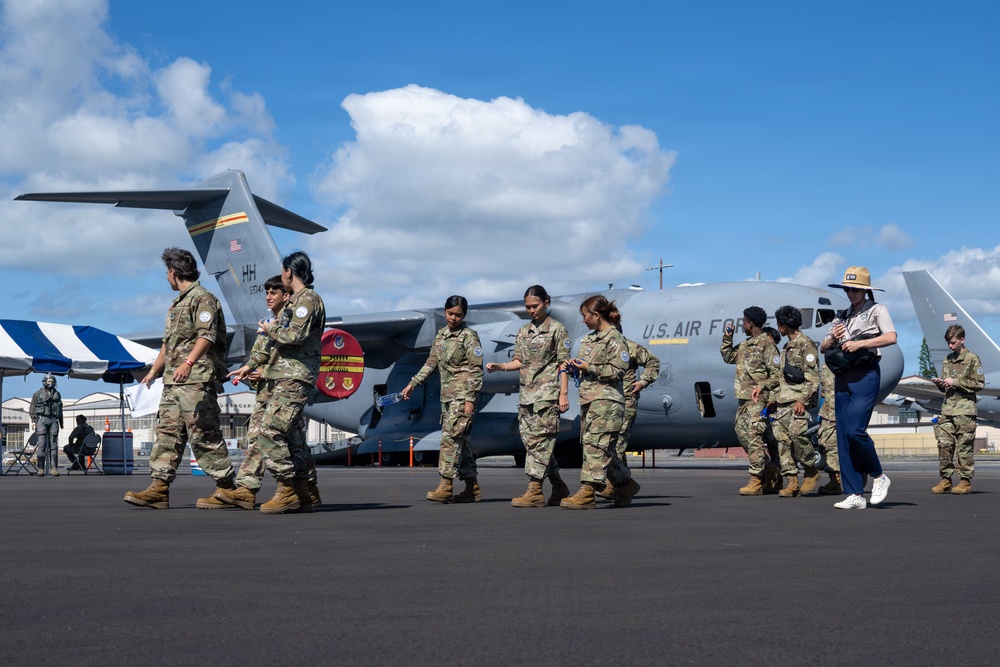 A group of military personnel in camouflage uniforms walk across an airfield tarmac in front of a U.S. Air Force C-17 Globemaster III transport aircraft. The aircraft bears the tail code 'HH' indicating it is assigned to Hawaii, along with unit patches and markings. A civilian guide wearing a ranger-style hat appears to be leading or accompanying the group, suggesting a tour or official visit.