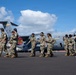 A group of military personnel in camouflage uniforms walk across an airfield tarmac in front of a U.S. Air Force C-17 Globemaster III transport aircraft. The aircraft bears the tail code 'HH' indicating it is assigned to Hawaii, along with unit patches and markings. A civilian guide wearing a ranger-style hat appears to be leading or accompanying the group, suggesting a tour or official visit.
