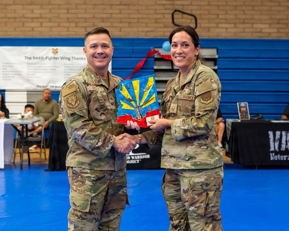 Two U.S. Air Force personnel in OCP (Operational Camouflage Pattern) uniforms are shaking hands while jointly holding a colorful plaque or award featuring what appears to be the Arizona state flag design with military aircraft. The setting is a gymnasium, with a banner in the background referencing the '944th Fighter Wing.' This appears to be a formal recognition or award ceremony.