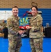 Two U.S. Air Force personnel in OCP (Operational Camouflage Pattern) uniforms are shaking hands while jointly holding a colorful plaque or award featuring what appears to be the Arizona state flag design with military aircraft. The setting is a gymnasium, with a banner in the background referencing the '944th Fighter Wing.' This appears to be a formal recognition or award ceremony.
