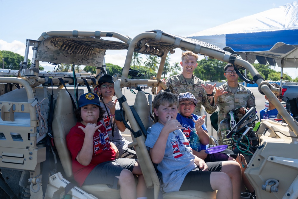 A group of children and two military personnel in camouflage uniforms pose together inside a tan-colored military tactical vehicle, likely during a public outreach or community event. Everyone in the photo is making the 'shaka' hand gesture, suggesting a Hawaiian location or cultural context. The setting appears to be an outdoor event with palm trees and a blue canopy visible in the background.