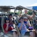 A group of children and two military personnel in camouflage uniforms pose together inside a tan-colored military tactical vehicle, likely during a public outreach or community event. Everyone in the photo is making the 'shaka' hand gesture, suggesting a Hawaiian location or cultural context. The setting appears to be an outdoor event with palm trees and a blue canopy visible in the background.