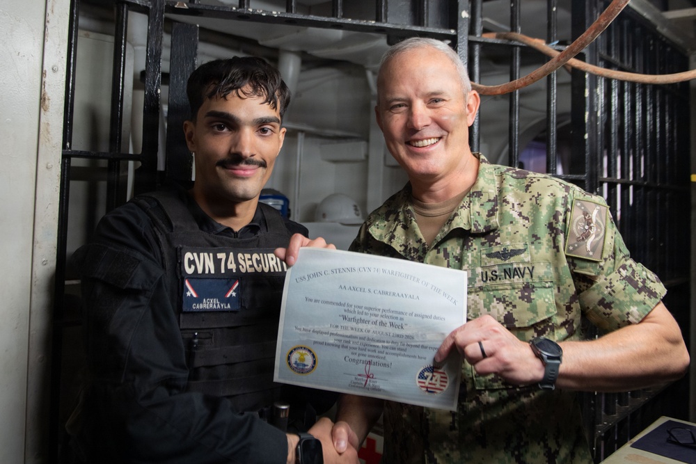 A U.S. Navy sailor assigned to CVN 74 Security is being presented with a 'Warfighter of the Week' certificate aboard USS John C. Stennis (CVN 74). The award recipient, identified on his name tape and the certificate as AA Axcel S. Cabreraayla, is posing with a senior U.S. Navy officer in NWU Type III camouflage uniform. Both individuals are smiling and holding the certificate together for the photo.