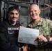 A U.S. Navy sailor assigned to CVN 74 Security is being presented with a 'Warfighter of the Week' certificate aboard USS John C. Stennis (CVN 74). The award recipient, identified on his name tape and the certificate as AA Axcel S. Cabreraayla, is posing with a senior U.S. Navy officer in NWU Type III camouflage uniform. Both individuals are smiling and holding the certificate together for the photo.