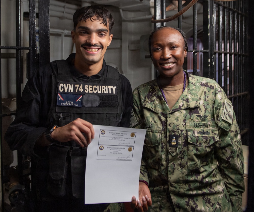 Two U.S. Navy personnel are posing together aboard what appears to be USS John C. Stennis (CVN-74). The individual on the left is wearing a CVN 74 Security vest with the name 'Cabreraayla' and is holding a 'Warfighter of the Week' certificate. The individual on the right is wearing standard U.S. Navy NWU Type III camouflage uniform with visible rank insignia and a name tag reading 'ILLIS'.
