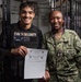 Two U.S. Navy personnel are posing together aboard what appears to be USS John C. Stennis (CVN-74). The individual on the left is wearing a CVN 74 Security vest with the name 'Cabreraayla' and is holding a 'Warfighter of the Week' certificate. The individual on the right is wearing standard U.S. Navy NWU Type III camouflage uniform with visible rank insignia and a name tag reading 'ILLIS'.