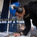 A U.S. Navy security personnel member is writing in a logbook at what appears to be a checkpoint or watch station. The individual is wearing a blue Navy hard hat with an official seal, hearing protection, safety glasses, and a black tactical security vest. An overlay text reads 'KEEPING SAILORS SAFE ONE LOG AT A TIME,' indicating this is likely an official U.S. Navy promotional or informational image.