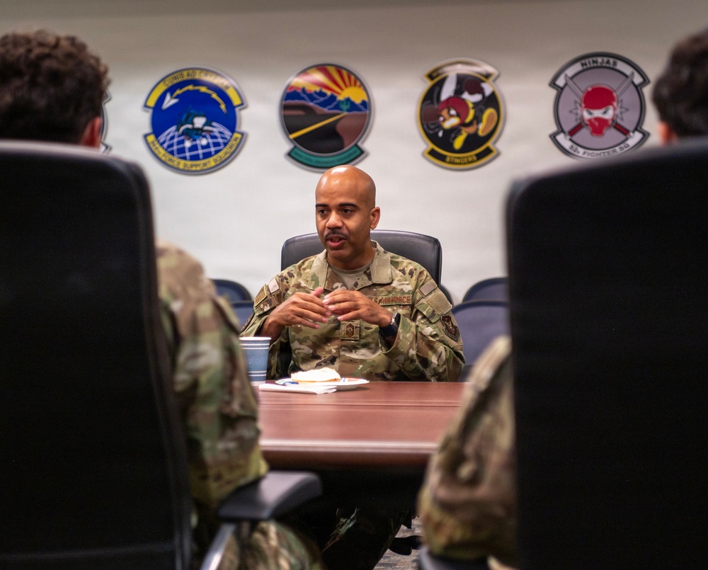 A U.S. Air Force senior enlisted member or officer is seated at a conference table, appearing to brief or address other uniformed personnel. The setting is a formal military meeting room with multiple squadron patches displayed on the wall behind the speaker. Two other uniformed individuals are visible from behind in the foreground, seated facing the speaker.