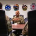 A U.S. Air Force senior enlisted member or officer is seated at a conference table, appearing to brief or address other uniformed personnel. The setting is a formal military meeting room with multiple squadron patches displayed on the wall behind the speaker. Two other uniformed individuals are visible from behind in the foreground, seated facing the speaker.
