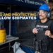 A security personnel member assigned to CVN 74 (USS John C. Stennis) is shown wearing a tactical vest labeled 'CVN 74 SECURITY', a blue hard hat, safety glasses, and hearing protection. The image appears to be an official U.S. Navy promotional or informational graphic with the overlay text 'SERVING AND PROTECTING HIS FELLOW SHIPMATES'. The setting appears to be aboard the aircraft carrier during a maintenance or industrial work period.
