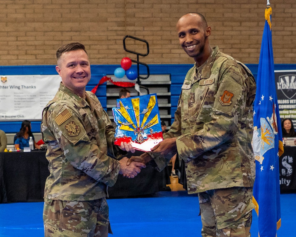 Two U.S. Air Force personnel in OCP (Operational Camouflage Pattern) uniforms are shaking hands during a formal presentation ceremony held in a gymnasium. One service member is presenting the other with a colorful plaque or award shaped like the state of Arizona with a sunburst design. The event appears to be a recognition or awards ceremony with banners, balloons, and an Air Force flag visible in the background.