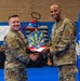 Two U.S. Air Force personnel in OCP (Operational Camouflage Pattern) uniforms are shaking hands during a formal presentation ceremony held in a gymnasium. One service member is presenting the other with a colorful plaque or award shaped like the state of Arizona with a sunburst design. The event appears to be a recognition or awards ceremony with banners, balloons, and an Air Force flag visible in the background.