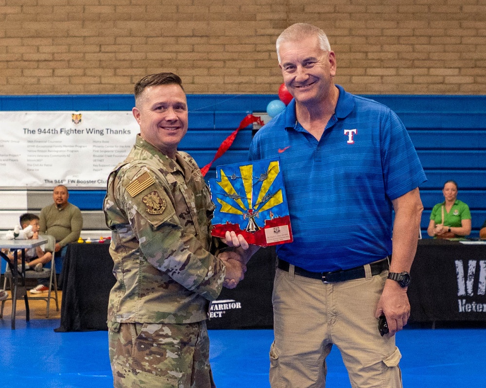 A military service member in Army Combat Uniform (ACU) is presenting an award or commemorative plaque to a civilian man wearing a Texas Rangers polo shirt. The ceremony appears to be taking place inside a gymnasium, with a banner visible in the background reading '944th Fighter Wing Thanks'. The award appears to be an Arizona-themed decorative piece featuring the state's sunburst design.