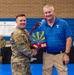 A military service member in Army Combat Uniform (ACU) is presenting an award or commemorative plaque to a civilian man wearing a Texas Rangers polo shirt. The ceremony appears to be taking place inside a gymnasium, with a banner visible in the background reading '944th Fighter Wing Thanks'. The award appears to be an Arizona-themed decorative piece featuring the state's sunburst design.