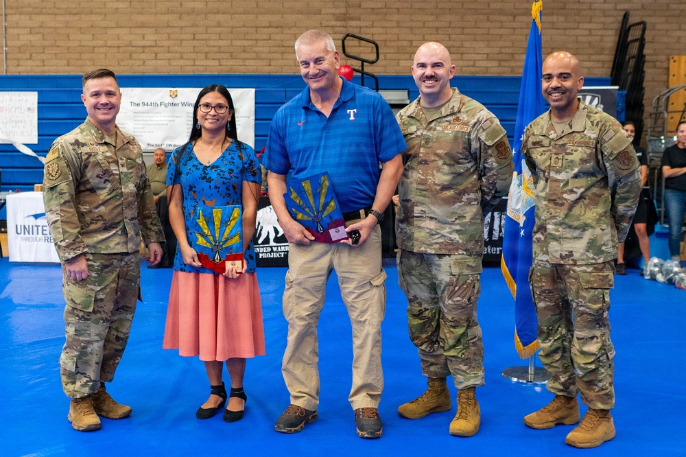 A group of five individuals pose for a formal photo in what appears to be a gymnasium or indoor facility. Four are wearing US Air Force OCP (Operational Camouflage Pattern) uniforms, while one civilian man in a blue polo shirt and one woman in a floral dress are also present. The civilian man is holding a decorative plaque featuring an Arizona state flag design, suggesting an award or recognition ceremony.